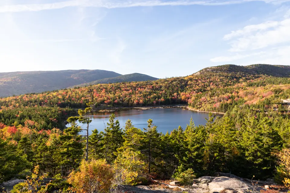 Blick von einer Granitkuppe über herbstlich rot, orange und gelb gefärbten Mischwald mit Nadelbäumen auf einen kleinen See. Dahinter liegen bewaldete Bergrücken unter hellblauem Himmel.