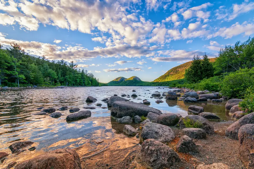 Ein stiller See mit großen gerundeten Steinen im flachen Uferwasser. Am Ende des Sees erheben sich zwei bewaldete, runde Berge unter einem Himmel mit Schleierwolken.