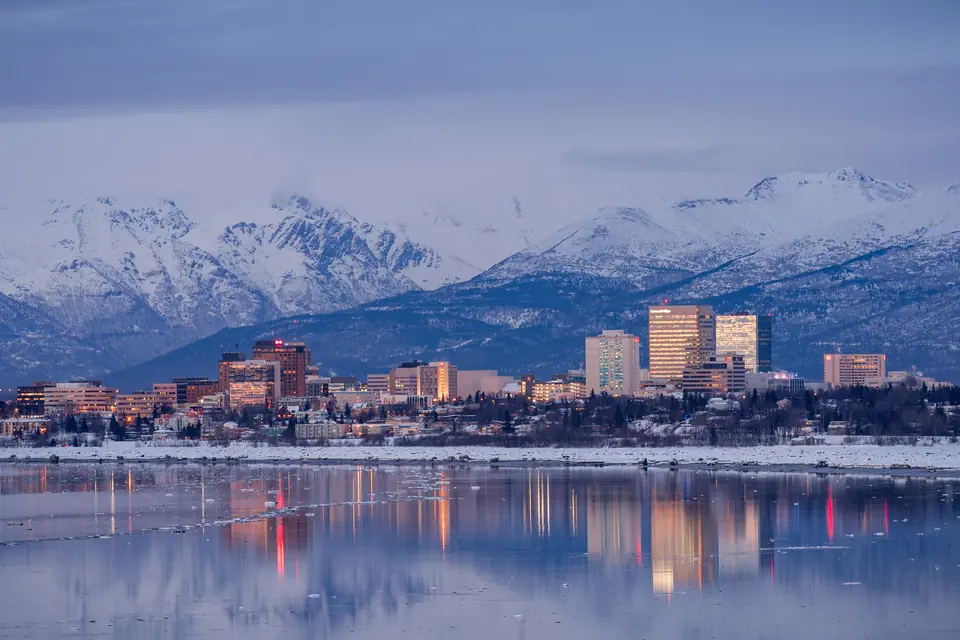 Hochhäuser einer Stadt am Ufer im Abendlicht, dahinter schneebedeckte Berge; die Lichter spiegeln sich im Wasser.