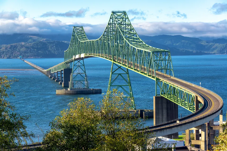 Grüne Stahlfachwerkbrücke mit geschwungener Auffahrt über breitem Wasser; dahinter bewaldete Hügel.