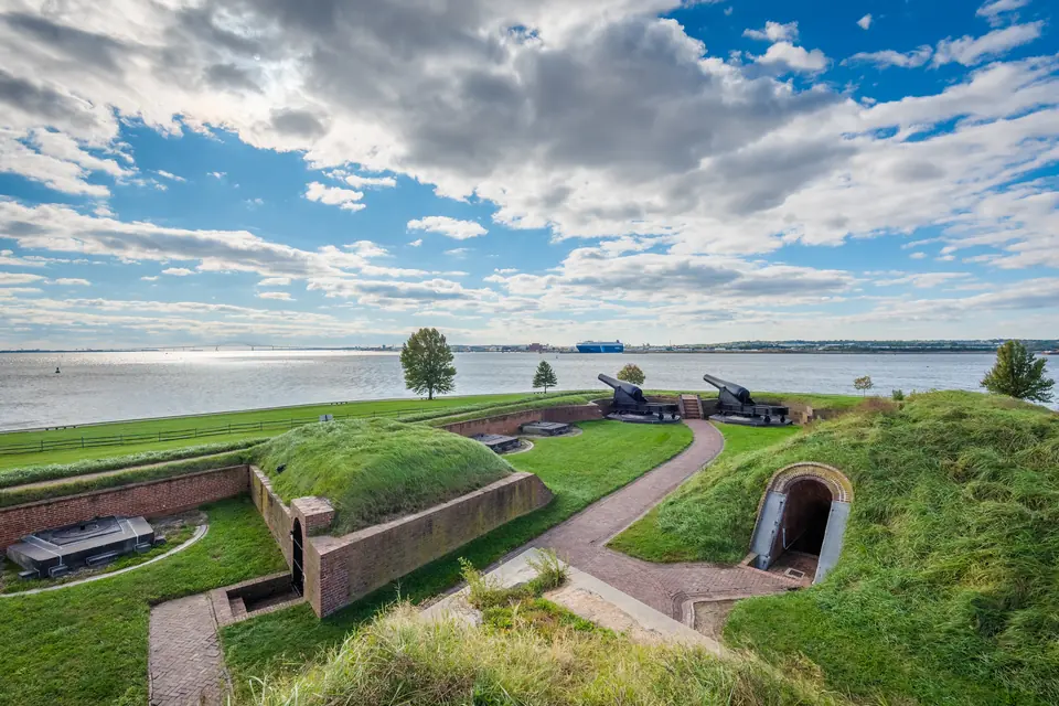 Grasbewachsene Wälle eines Sternforts mit gemauertem Durchgang und Kanonen, dahinter eine weite Wasserfläche unter Wolken.