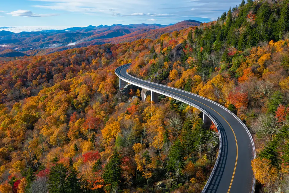 Luftbild einer geschwungenen Straßenbrücke auf Betonpfeilern, die durch rot und gelb gefärbten Herbstwald am Berghang führt, dahinter Bergketten.