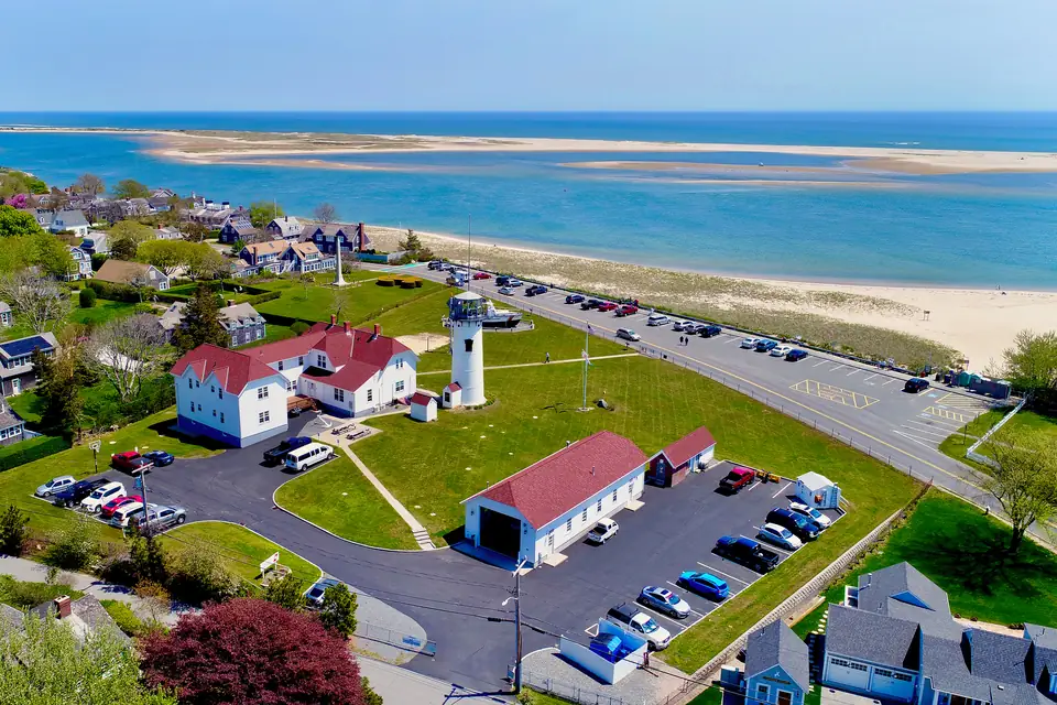 Luftbild eines weißen Leuchtturms neben weißen Gebäuden mit roten Dächern und einem Parkplatz. Dahinter liegen ein Strand, eine lange Sandbank und das offene Meer.