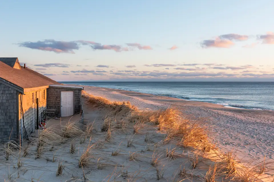 Eine mit grauen Holzschindeln verkleidete Hütte zwischen Dünengras am Rand eines breiten Sandstrands. Rechts liegt das Meer, der Himmel ist abendlich hell.