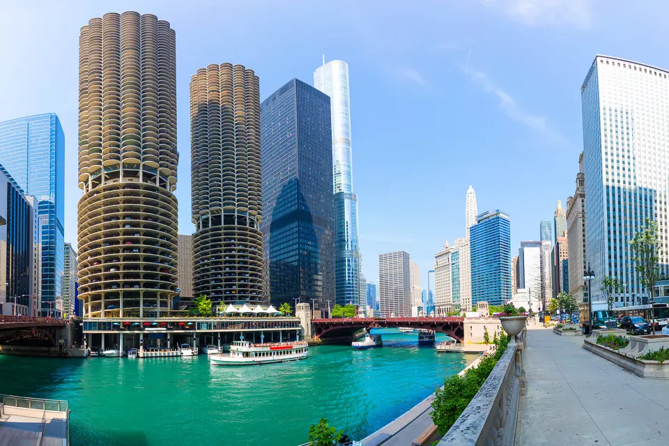 Die beiden runden Türme von Marina City mit ihren Balkonreihen am türkisgrünen Chicago River. Am Fuß der Türme liegen Boote in der Marina, auf dem Fluss fährt ein Ausflugsboot. Rechts verläuft ein Uferweg mit Pflanzkübeln, im Hintergrund stehen Hochhäuser und eine rote Brücke.