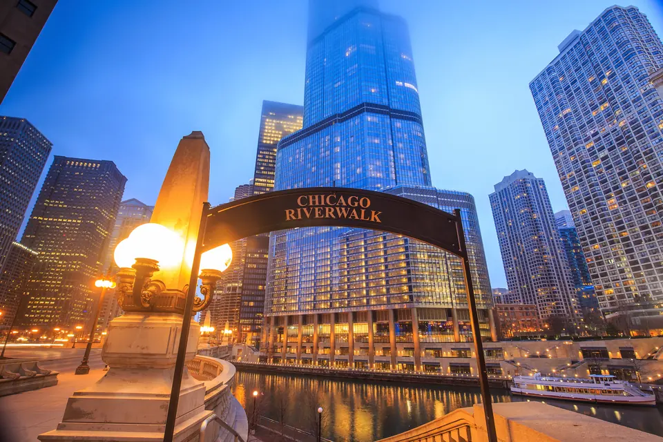 Ein Schildbogen mit der Aufschrift Chicago Riverwalk in der Abenddämmerung, links eine leuchtende Laterne auf einem steinernen Pfeiler. Dahinter liegen der beleuchtete Fluss mit einem Ausflugsboot und ein gläsernes Hochhaus, dessen Spitze im Nebel verschwindet.