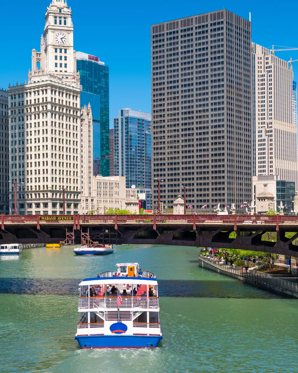 Ein weiß-blaues Ausflugsboot auf dem grünen Chicago River vor einer roten Stahlbrücke mit der Aufschrift Wabash Avenue; dahinter links der weiße Uhrturm des Wrigley Building, rechts ein Uferweg mit Fußgängern.