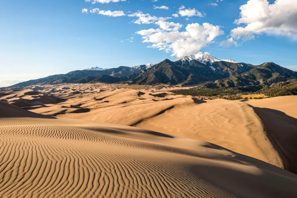 Sanddünen mit geriffelter Oberfläche vor einer Bergkette mit schneebedeckten Gipfeln unter blauem Himmel.