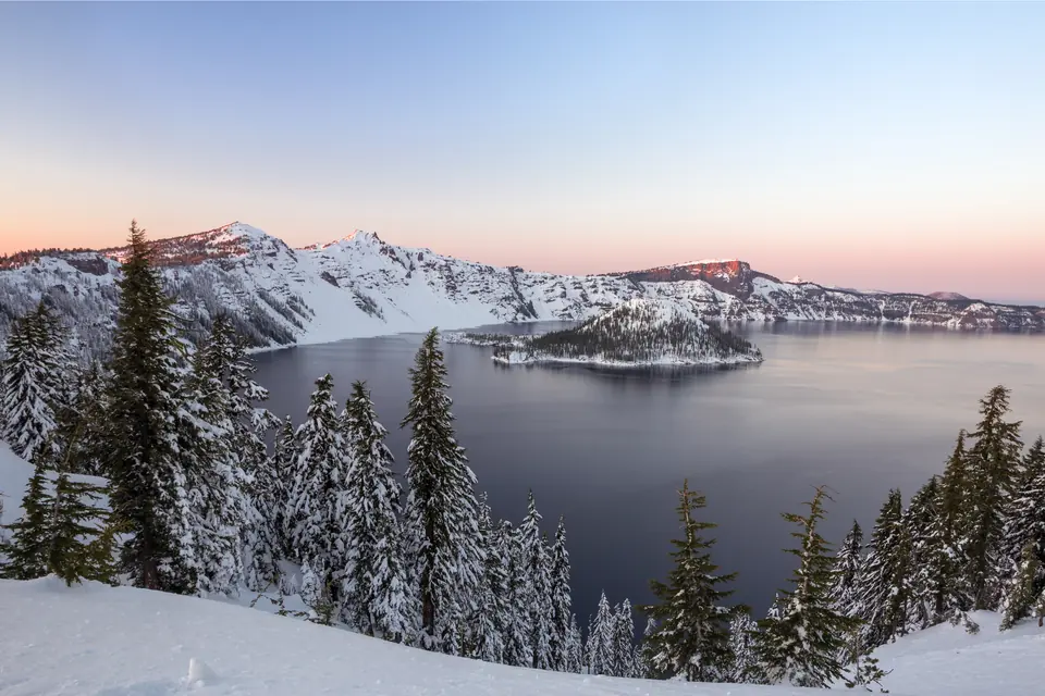 Verschneite Kesselwände umschließen einen ruhigen See mit einer schneebedeckten Insel, davor tief verschneite Nadelbäume im Abendlicht.