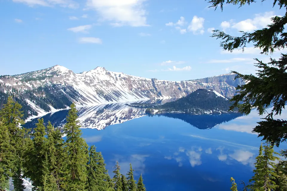 Steile, teils verschneite Felswände mit Gesteinslagen spiegeln sich im glatten blauen See, rechts davor eine bewaldete Insel.