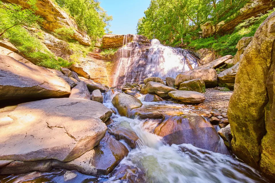 Wasserfall, der über eine überstehende Sandsteinkante und eine dünn geschichtete Felswand fällt, davor ein Bach zwischen großen Felsblöcken und Laubbäume an den Hängen.