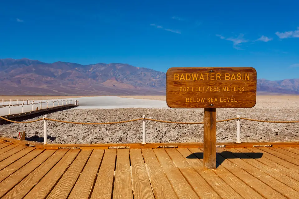 Ein Holzsteg mit einem braunen Schild „Badwater Basin, 282 feet below sea level“ (rund 86 Meter unter dem Meeresspiegel); dahinter die helle Salzfläche und eine Bergkette unter blauem Himmel.