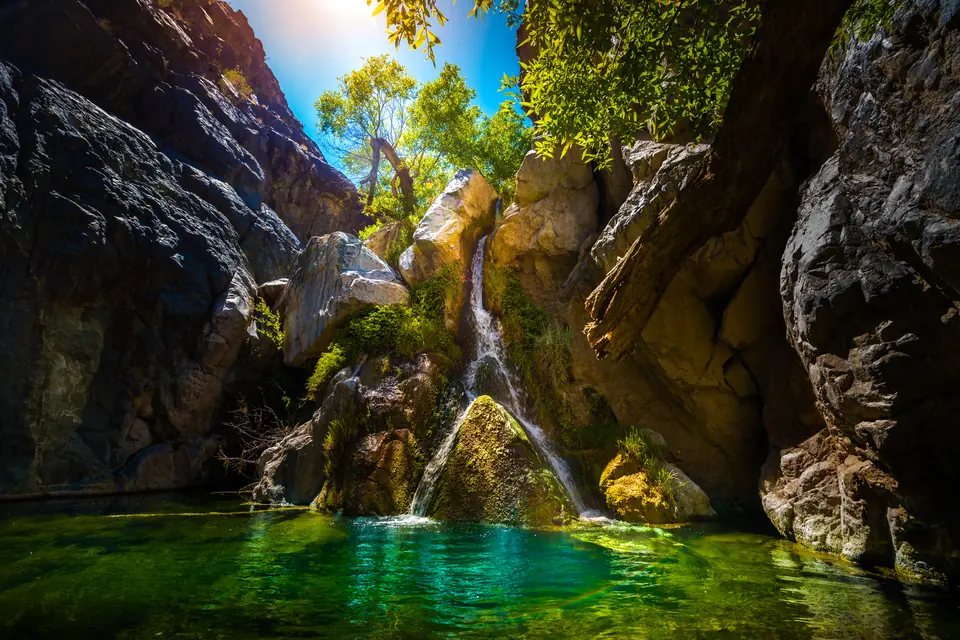 Schmaler Wasserfall zwischen Felsblöcken, der in ein klares Becken fällt, oben grüne Bäume.