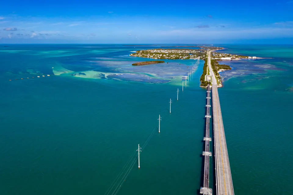 Luftbild: eine breite Straßenbrücke führt neben einer schmaleren älteren Brücke auf Pfeilern über türkisfarbenes Flachwasser zu einer Insel mit Häusern, daneben eine Reihe Strommasten.