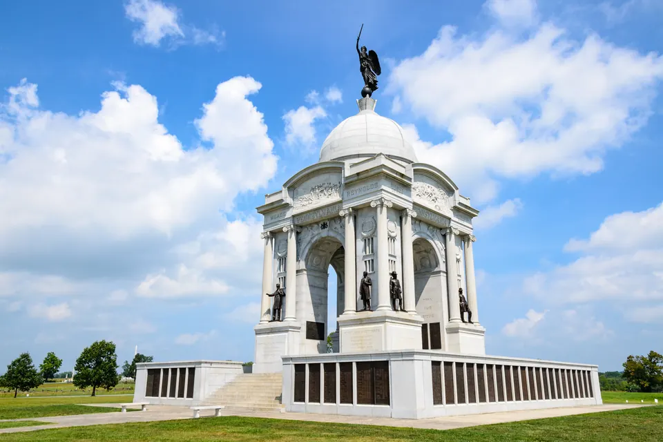 Ein weißes Denkmal mit Kuppel, Siegesfigur auf der Spitze und Bronzestatuen zwischen den Säulen, am Sockel Reihen von Bronzetafeln, darüber Sommerwolken.