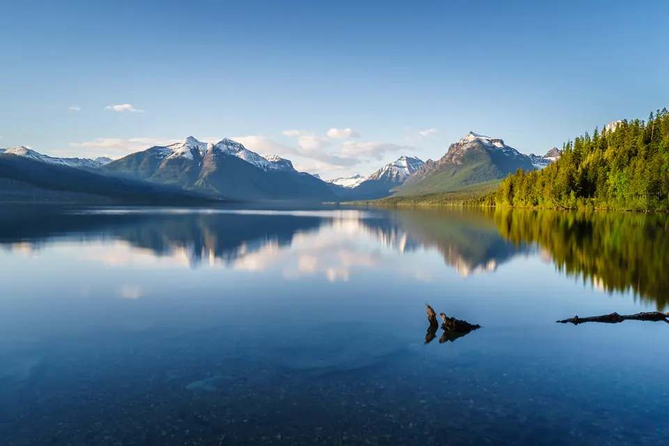 Ruhiges Seewasser spiegelt bewaldete Ufer und schneebedeckte Berge; im Vordergrund liegen einzelne Holzstücke.