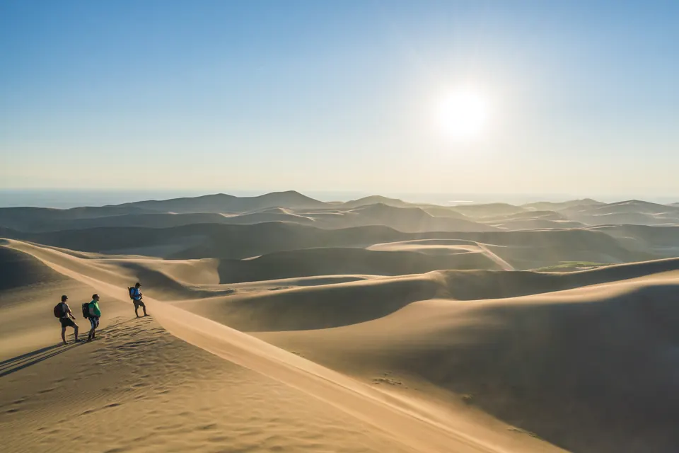 Drei Wanderer mit Rucksäcken auf einem Dünengrat im Gegenlicht, dahinter weitere Dünenkämme bis zum Horizont.