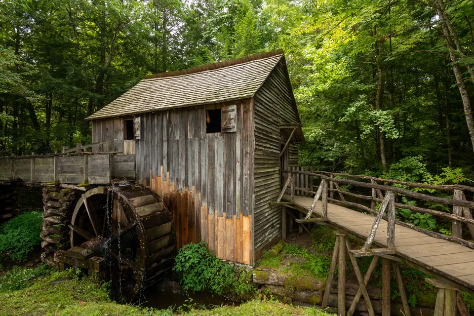 Hölzerne Mühle mit großem Wasserrad und einem Holzsteg zwischen grünen Bäumen.