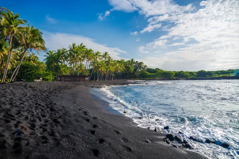 Ein Strand aus schwarzem Sand mit Fußspuren, an dem Wellen auslaufen; dahinter ein Saum aus Kokospalmen unter weißen Wolken.
