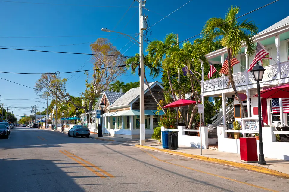 Eine sonnige Straße in Key West mit weißen Holzhäusern, Veranden, Palmen und amerikanischen Flaggen, im Hintergrund parkende Autos.