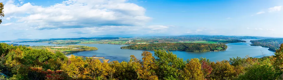 Weiter Blick über Bäume im Herbstlaub auf den Lake Champlain mit bewaldeten Inseln und flachem Ufer unter blauem Himmel.