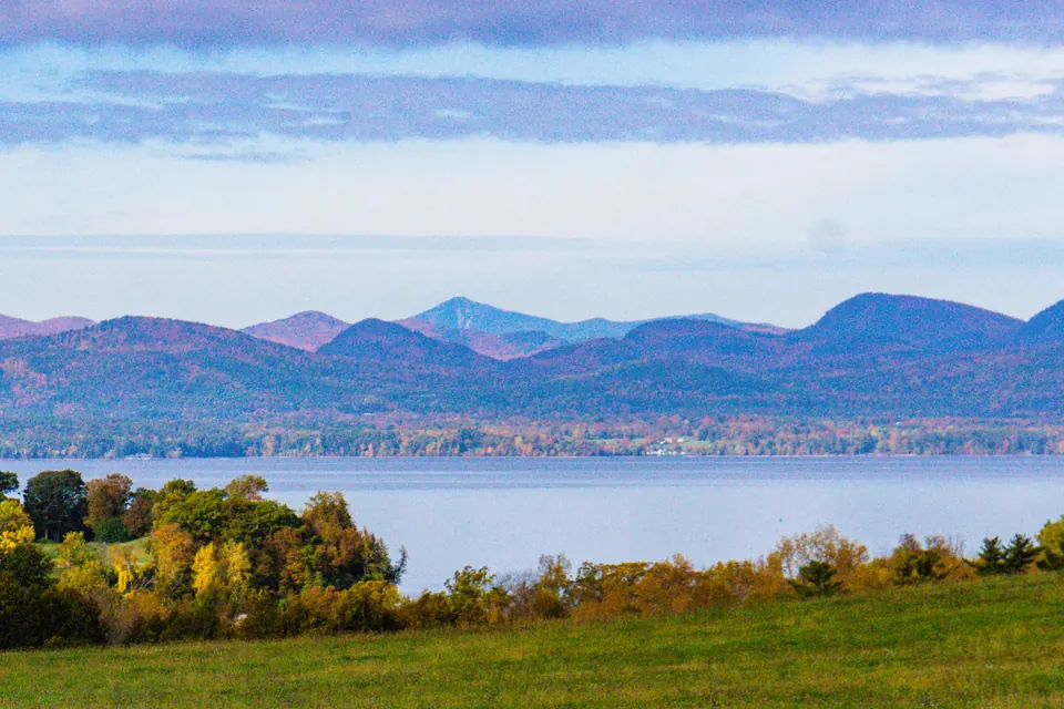 Eine Wiese und Bäume im Herbstlaub vor dem Lake Champlain, dahinter blaue Bergketten unter einem hellen Himmel.