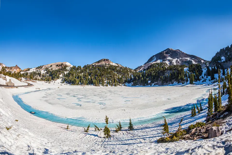 Ein zugefrorener, verschneiter Bergsee mit einem schmalen Streifen türkisfarbenen Wassers am Ufer, ringsum Schnee und Nadelbäume, dahinter Berggipfel mit Schneefeldern unter blauem Himmel.