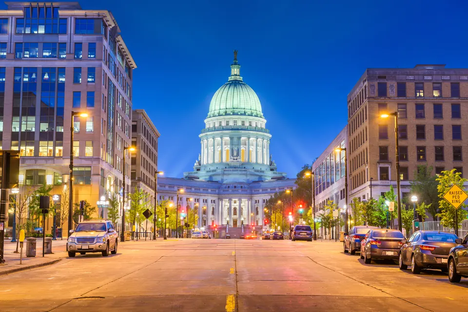 Die beleuchtete Kuppel des Wisconsin State Capitol steht am Ende einer Straße zwischen Häusern und Straßenlaternen.
