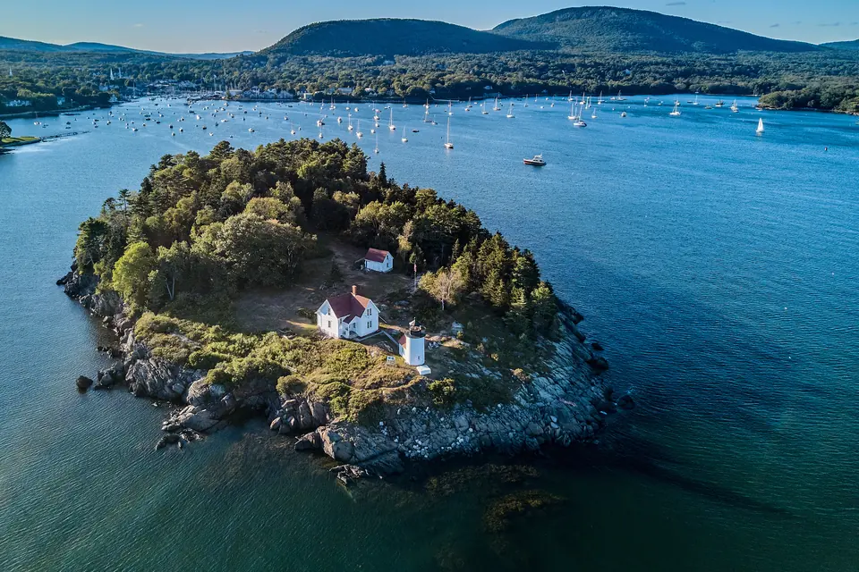 Luftbild einer bewaldeten Felsinsel mit weißem Leuchtturm und Wärterhaus mit rotem Dach. Dahinter liegt eine Bucht voller Segelboote, am Ufer ein Ort vor bewaldeten Bergen.