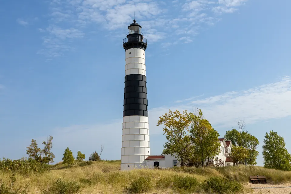 Schwarz-weiß gestreifter Leuchtturm mit Wärterhaus zwischen Dünengras und Bäumen unter blauem Himmel.