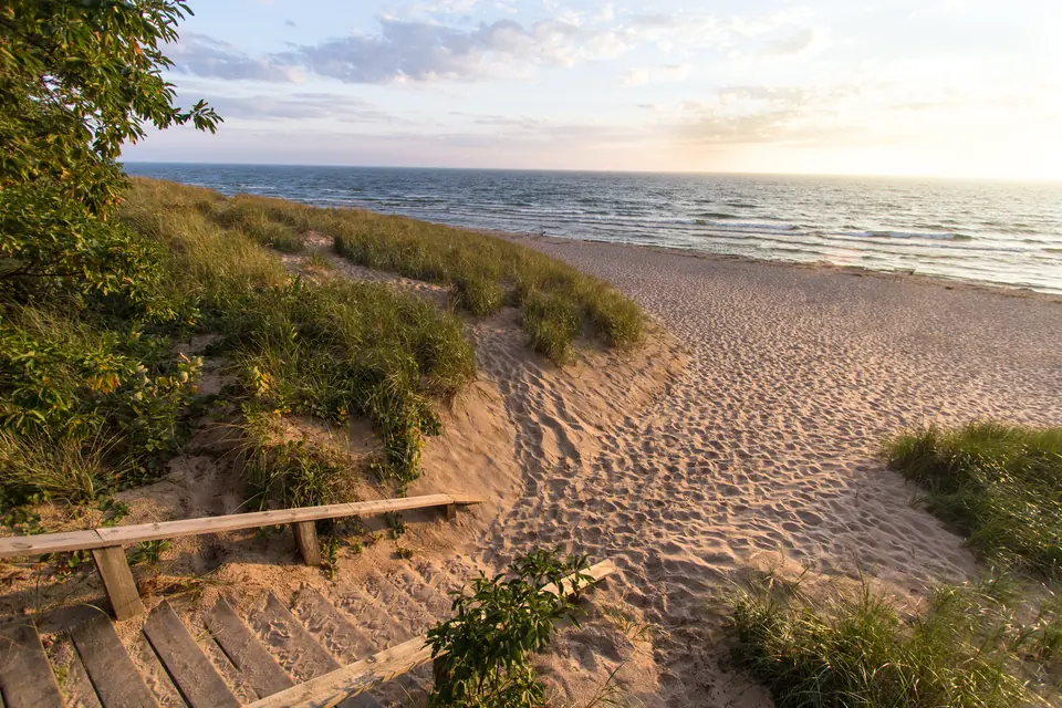 Holzstufen und ein Sandweg führen zwischen bewachsenen Dünen hinunter zu einem breiten Strand am See.