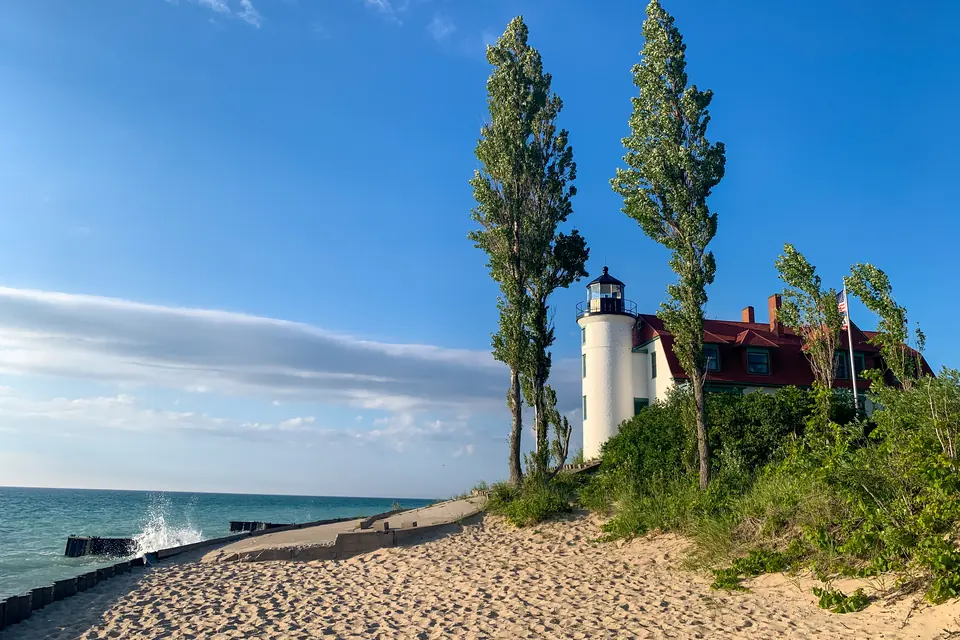 Weißer Leuchtturm mit rotem Dach hinter zwei hohen Pappeln, davor ein Sandstrand mit Uferbefestigung am See.