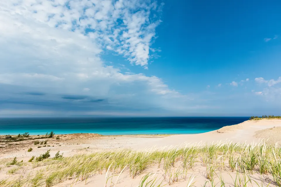 Heller Sand mit Dünengras, dahinter türkisfarbenes Wasser des Lake Michigan unter blauem Himmel mit Wolken.
