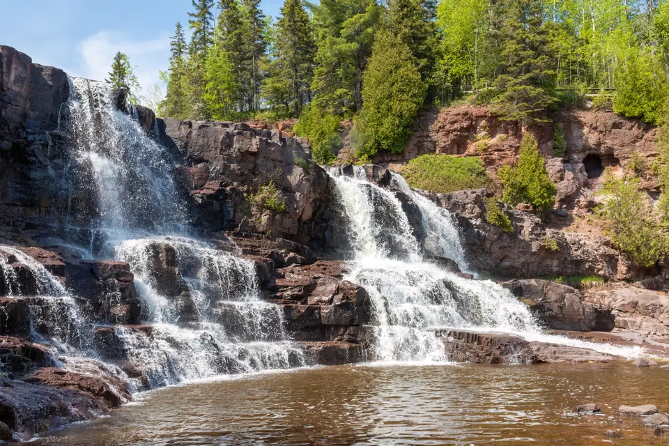 Wasser fällt über mehrere Stufen aus dunklem Fels in ein Becken, darüber Laub- und Nadelwald.