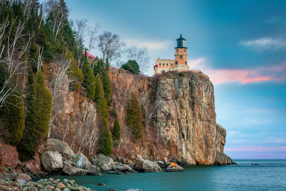 Ein Leuchtturm mit gelbem Backsteinturm steht auf einer steilen Felsklippe über dem See, unten Ufersteine.