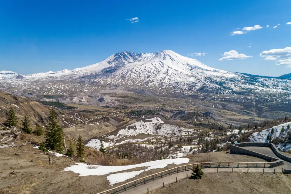 Der schneebedeckte Mount St. Helens mit seinem nach Norden offenen Krater über einem kahlen, teils verschneiten Tal. Im Vordergrund ein Weg mit Geländer an einem Aussichtspunkt.