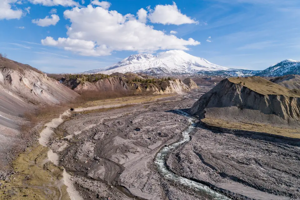 Ein Fluss windet sich durch ein breites Tal aus grauem Geröll zwischen abgetragenen Hängen; im Hintergrund der schneebedeckte Mount St. Helens.