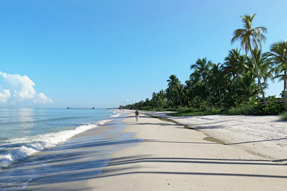 Breiter, heller Sandstrand an ruhigem Wasser mit flachen Wellen, rechts Palmen, ein einzelner Spaziergänger.