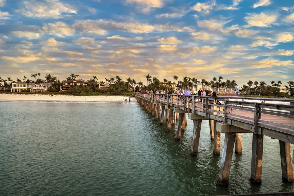 Ein langer hölzerner Pier auf Pfählen führt vom Strand ins ruhige Wasser, auf ihm gehen einige Menschen. Am Ufer stehen Palmen, darüber ziehen Schäfchenwolken.