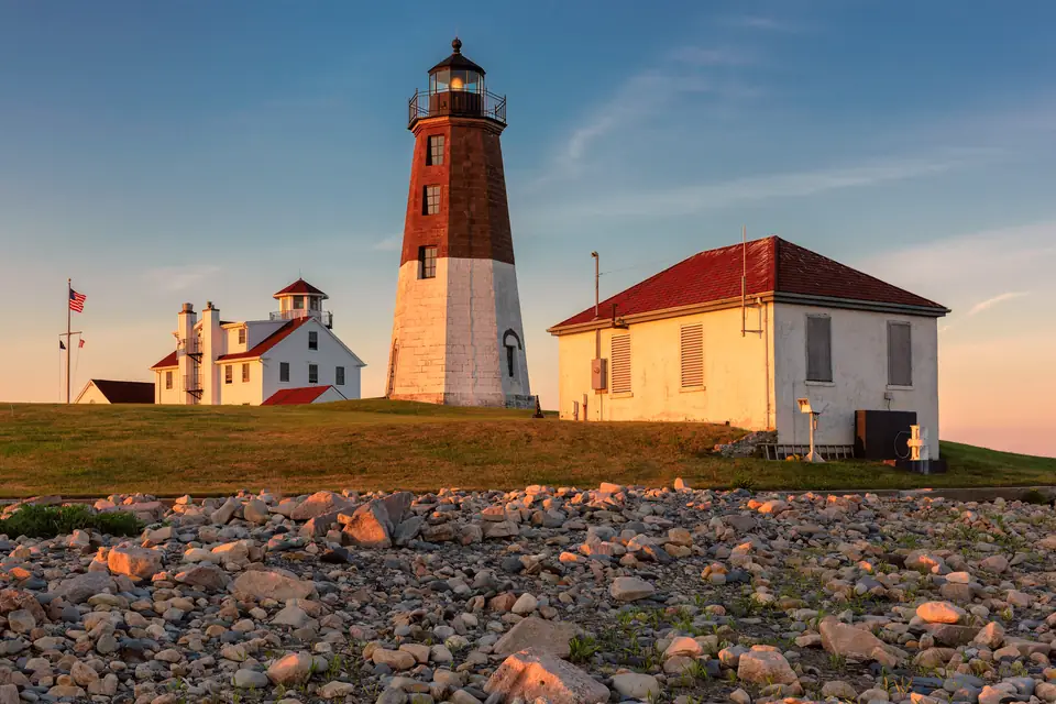 Achteckiger Leuchtturm, unten weiß, oben rotbraun, daneben Nebengebäude mit roten Dächern, im Vordergrund ein Steinstrand im Abendlicht.