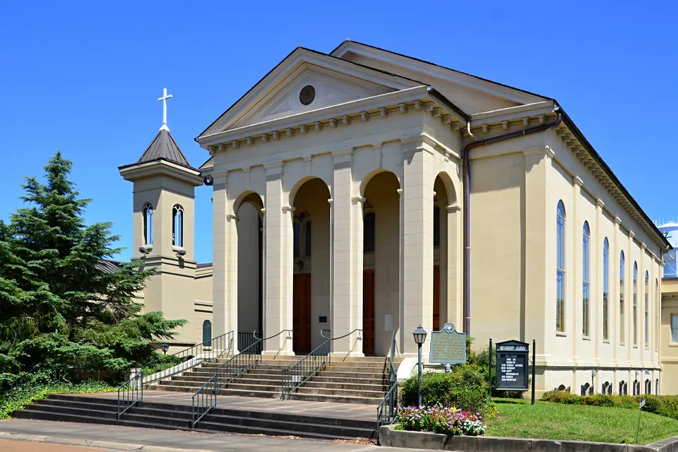 Hell gestrichene Kirche mit Bogenhalle, Freitreppe und Glockenturm unter blauem Himmel.