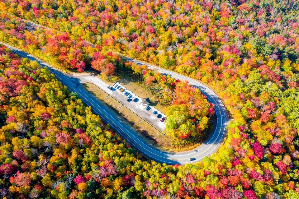 Luftbild einer Haarnadelkurve, in deren Innenseite ein Parkplatz liegt, umgeben von dichtem Laubwald.