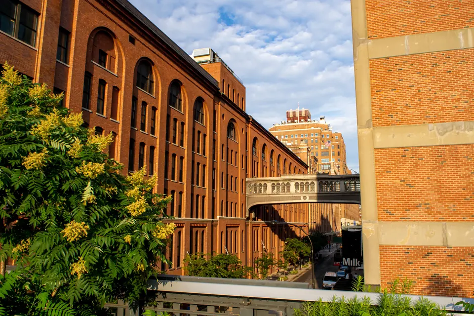 Blick von der High Line in eine Straße mit langen Backsteinbauten und hohen Bogenfenstern, die eine Fußgängerbrücke über die Straße verbindet.