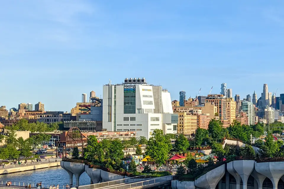 Das helle Whitney Museum mit dem Schriftzug WHITNEY an der Fassade; davor eine begrünte Anlage auf tulpenförmigen Pfeilern am Hudson, dahinter die Stadt.