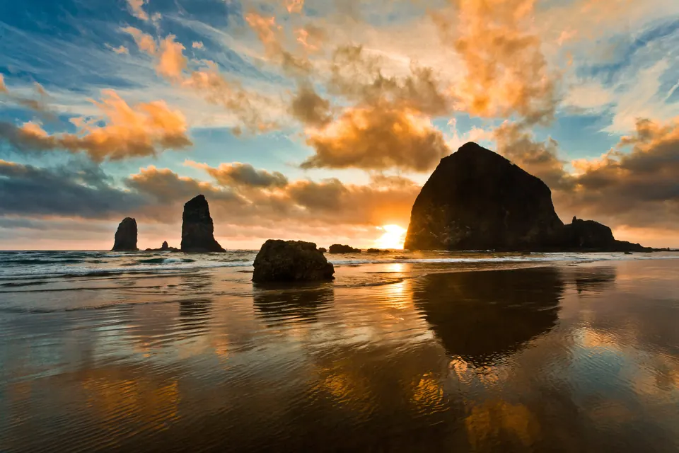 Großer dunkler Felsen und zwei schmale Felsnadeln im Meer vor einem Sandstrand, orange beleuchtete Wolken und Spiegelungen im nassen Sand.