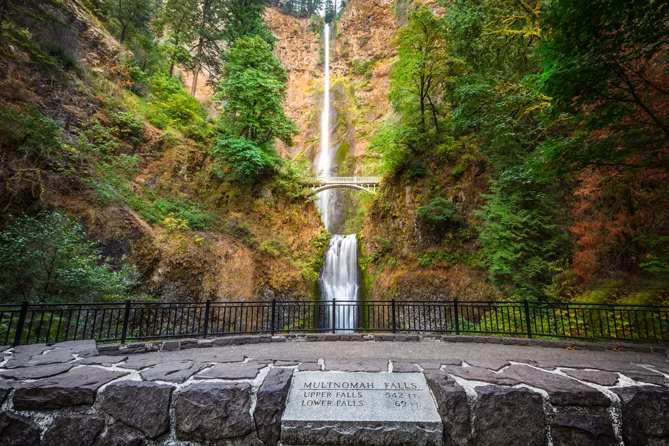 Hoher, zweistufiger Wasserfall in einer bewachsenen Felswand, davor eine Steinmauer mit eingelassener Inschriftentafel und ein Geländer.