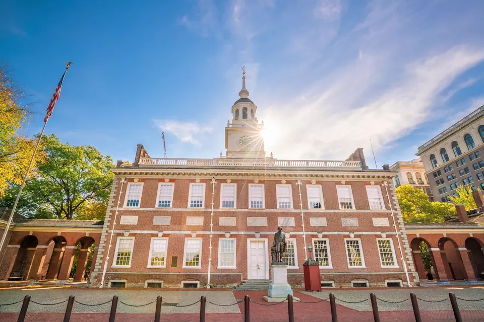 Die Independence Hall, ein langer Backsteinbau mit weißen Fensterrahmen und einem weißen Turm, im Gegenlicht. Davor steht eine Statue auf einem Sockel, links weht eine Flagge.