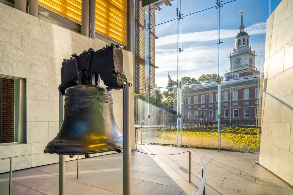 Die Liberty Bell hängt an einem dunklen Holzjoch in einem hellen Raum hinter einer niedrigen Absperrung. Durch eine hohe Glaswand ist die Independence Hall mit ihrem weißen Glockenturm zu sehen.