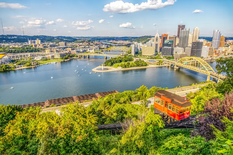 Blick von einem grünen Hang auf die Spitze der Innenstadt von Pittsburgh. Dort treffen zwei Flüsse zusammen; an der Spitze liegt ein Park, dahinter stehen Hochhäuser und gelbe Brücken. Vorn steht ein roter Wagen einer Standseilbahn am Hang.