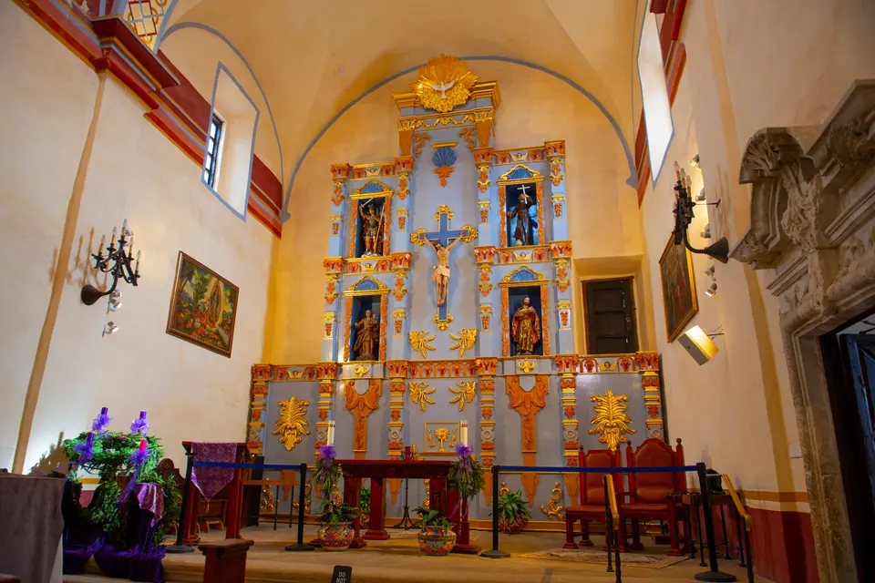 Innenraum einer Missionskirche mit farbig gefasstem Altaraufsatz, Kruzifix und Heiligenfiguren, davor Altar und Stuhlreihen.